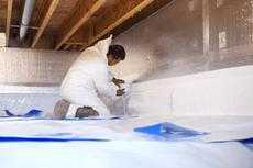 A person in protective gear applies insulation in a crawl space with wooden beams.