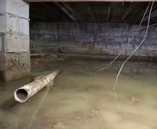 Flooded crawl space under a house with standing water, mud, debris, and exposed pipe.