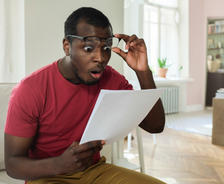 A surprised man in glasses reads a paper while sitting on a couch in a bright living room.