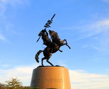 Bronze horse and rider statue holding a spear, atop a pedestal labeled “UNCONQUERED” against a blue sky.