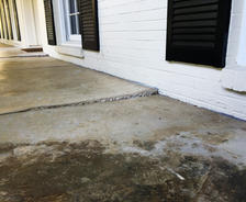 Concrete porch with a noticeable crack and an uneven surface, ready for expert concrete patio repair, near a white brick house with black shutters.