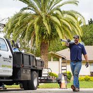 A worker carries a pipe near a truck on a sunny day, with a palm tree and a house in the background.