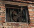 A crawl space fan, old and rusty, sits securely behind bars in a brick wall window with a weathered wood frame.