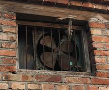 A crawl space fan, old and rusty, sits securely behind bars in a brick wall window with a weathered wood frame.