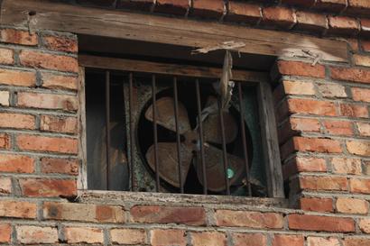A crawl space fan, old and rusty, sits securely behind bars in a brick wall window with a weathered wood frame.