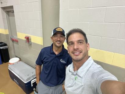 Two men taking a selfie indoors near a cooler and fire alarm, both wearing casual shirts and smiling.