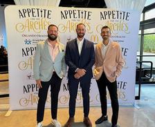 Three men in suits smiling in front of a backdrop at an event titled "Appetite for the Arches.