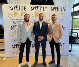 Three men in suits smiling in front of a backdrop at an event titled "Appetite for the Arches.