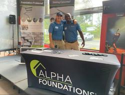 Two smiling people standing behind an "Alpha Foundations" table at an indoor event.