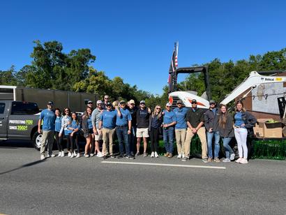 A group of people posing next to a truck and construction vehicle on a sunny day.