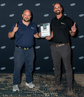 Two men smiling and holding an award, standing in front of a backdrop with repeated text.