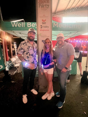 Three people smiling at a nighttime outdoor event under a tent with sponsor signs in the background.