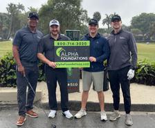 Four men smiling and holding an Alpha Foundations sign on a rainy golf course.
