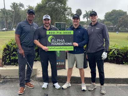 Four men smiling and holding an Alpha Foundations sign on a rainy golf course.
