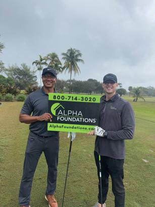 Two men holding an "Alpha Foundations" sign on a golf course, with palm trees and cloudy skies in the background.