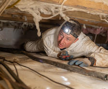 In a confined space, a person in white coveralls and headlamp inspects for mold, surrounded by insulation and cables.