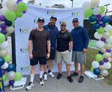 Four men stand smiling in front of a "Hearts 4 Minds" banner, surrounded by a balloon arch.
