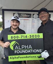 Two men smile while holding an Alpha Foundations sign at a golf event, with a scoreboard in the background.