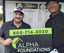 Two men smile while holding an Alpha Foundations sign at a golf event, with a scoreboard in the background.
