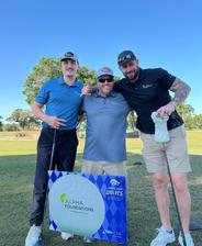 Three men smiling on a golf course, standing behind a sign for Alpha Foundations and Jacksonville Wolves.