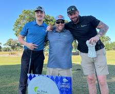 Three men smiling on a golf course, standing behind a sign for Alpha Foundations and Jacksonville Wolves.