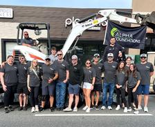 Group of people in matching shirts posing by a small excavator, with an Alpha Foundations banner in the background.
