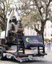Excavator with Alpha Foundations banner on a parade float, surrounded by spectators and trees.