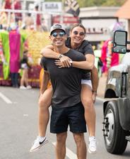 Man giving a piggyback ride to a woman at a festive outdoor event with colorful floats.