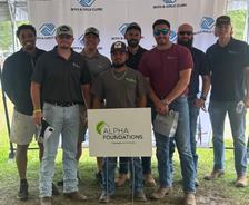 Group of men posing under a tent with an "Alpha Foundations" sign at a Boys & Girls Clubs event.