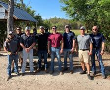 Eight men stand outdoors in casual attire, some wearing caps and sunglasses, with trees and a small structure in the background.