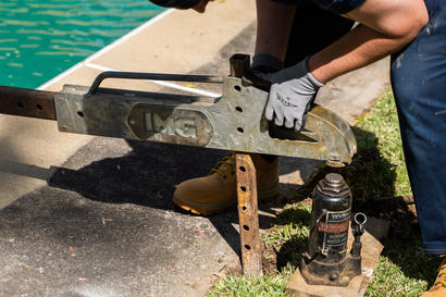 Person using a hydraulic jack for pool deck repair, lifting a metal structure near the poolside.