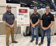 Three men stand next to a sponsor board inside a showroom with tile flooring and displays in the background.