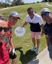 Four men smiling on a golf course, one holding a sign that reads "Alpha Foundations.