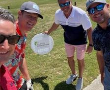 Four men smiling on a golf course, one holding a sign that reads "Alpha Foundations.