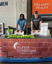Two men stand behind an Alpha Foundations booth with green giveaways at an indoor event.