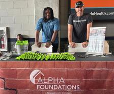 Two men stand behind an Alpha Foundations booth with green giveaways at an indoor event.