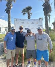 Four men in casual clothes pose together outdoors by palm trees and a sponsor banner on a sunny day.