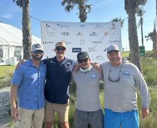 Four men in casual clothes pose together outdoors by palm trees and a sponsor banner on a sunny day.