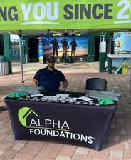 Man sitting at an Alpha Foundations booth with brochures and swag under a green promotional tent.