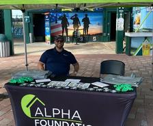 Man sitting at an Alpha Foundations booth with brochures and swag under a green promotional tent.