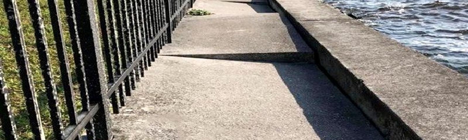 A narrow sidewalk runs beside a metal fence and waterfront, with uneven, broken concrete sections.