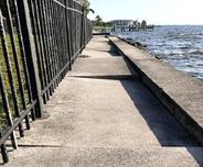 A narrow sidewalk runs beside a metal fence and waterfront, with uneven, broken concrete sections.