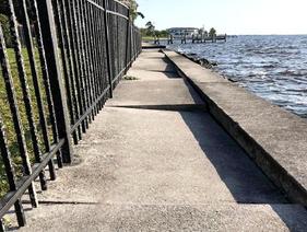 A narrow sidewalk runs beside a metal fence and waterfront, with uneven, broken concrete sections.