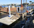Workers construct a seawall along a waterfront, with equipment, pilings, and homes visible in the background.