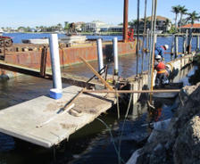 Workers construct a seawall along a waterfront, with equipment, pilings, and homes visible in the background.