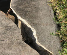 A large crack splits a concrete sidewalk beside grass and water.