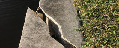 A large crack splits a concrete sidewalk beside grass and water.