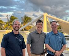 Three men smiling outdoors, standing in front of palm trees and a white structure under a cloudy sky.