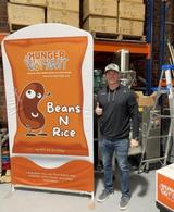 Man giving thumbs up next to a “Hunger Fight Beans N Rice” display in a warehouse setting.