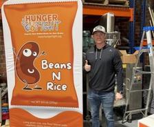 Man giving thumbs up next to a “Hunger Fight Beans N Rice” display in a warehouse setting.